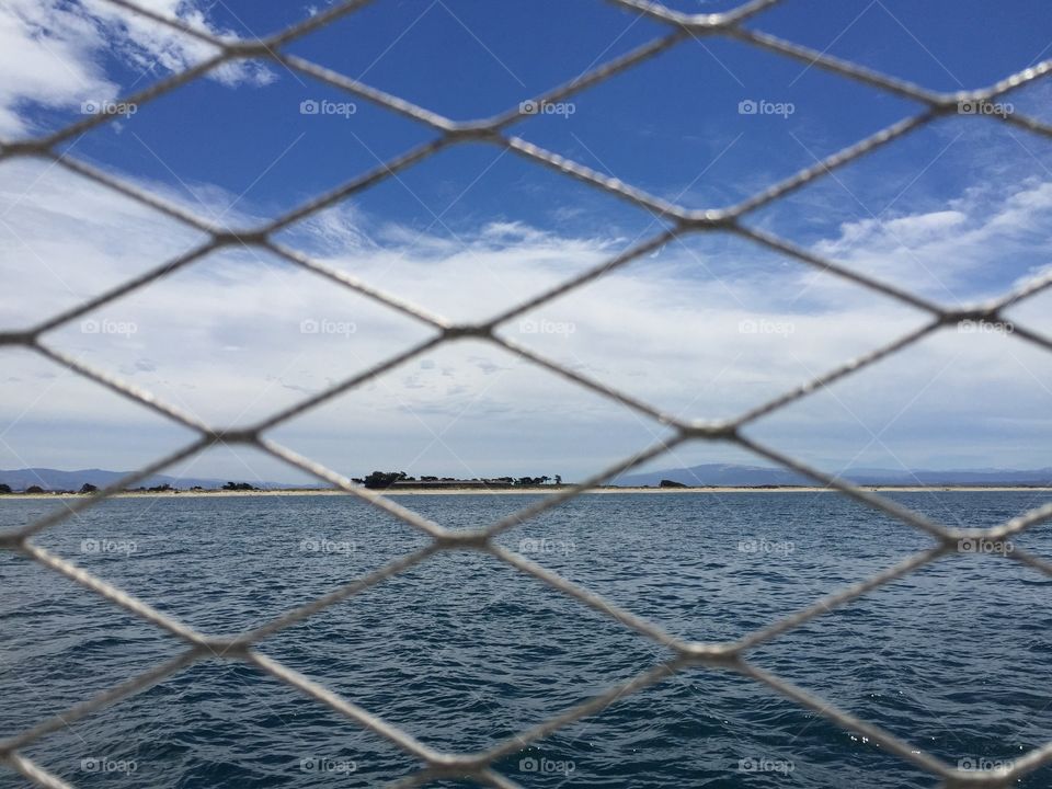 View of ocean water and coast, through a mesh of railing on boat.
