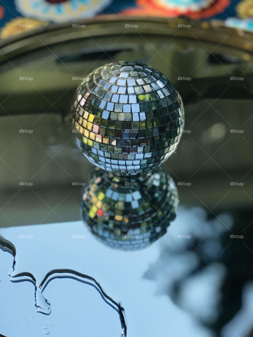 Filtered sunlight shines on a disco ball resting on water on a mirror. Tree branches  & blue sky are reflected in the mirror. The surrounding landscape is reflected in the tiny glass panes of the ball creating a symmetrical kaleidoscope of colours.