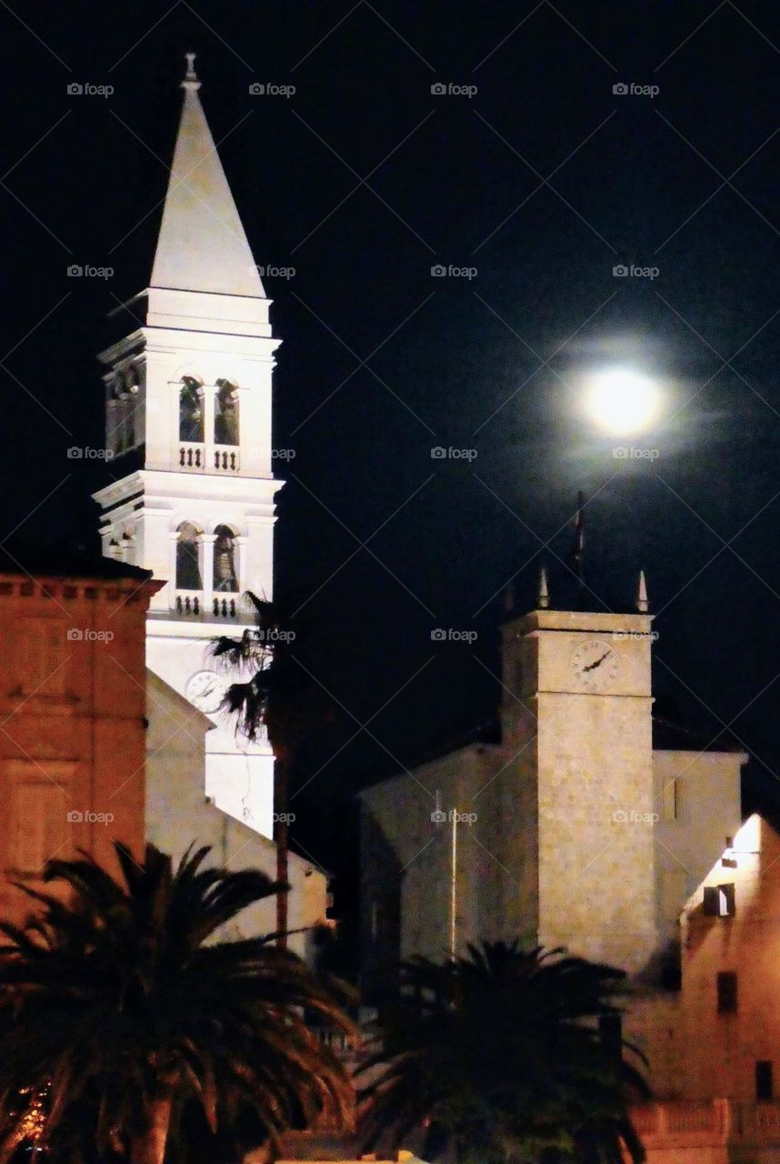 Full moon reflecting on a Venetian tower on the left and a clock tower on the right with silhouettes of date palms in the foreground