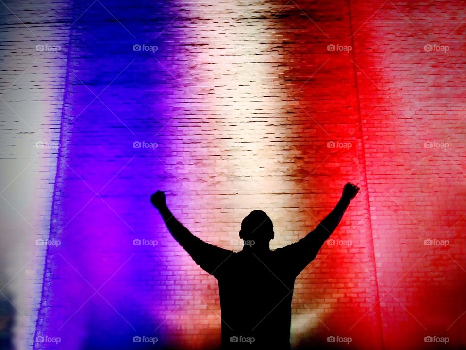 Silhouette of man with his arms up in the air in front of red white and blue lighted backdrop