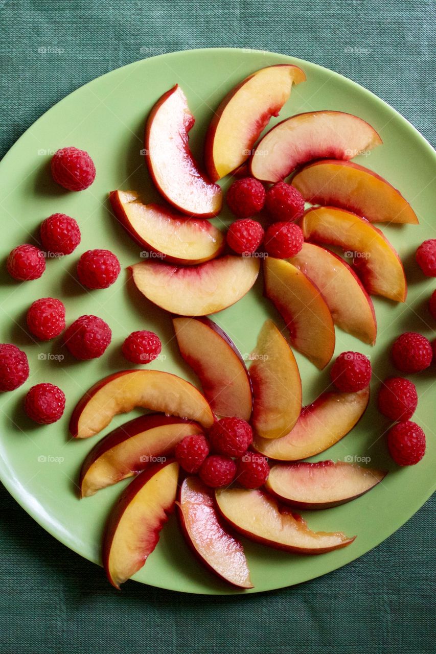 Flat lay of a decorative arrangement of fresh nectarine slices and raspberries on a green plate with green background 