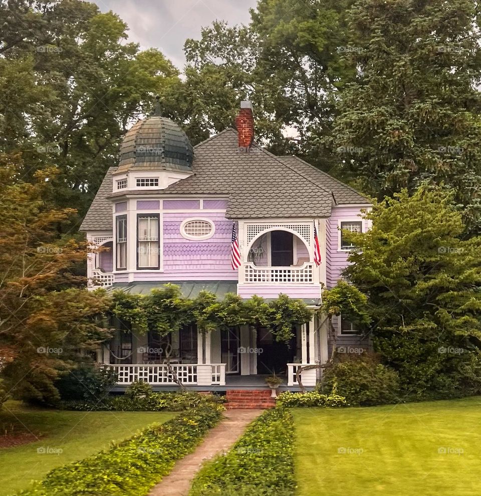 Two-story purple Victorian house with unique roof design surrounded by shrubs and a path leading towards the front porch through a manicured lawn
