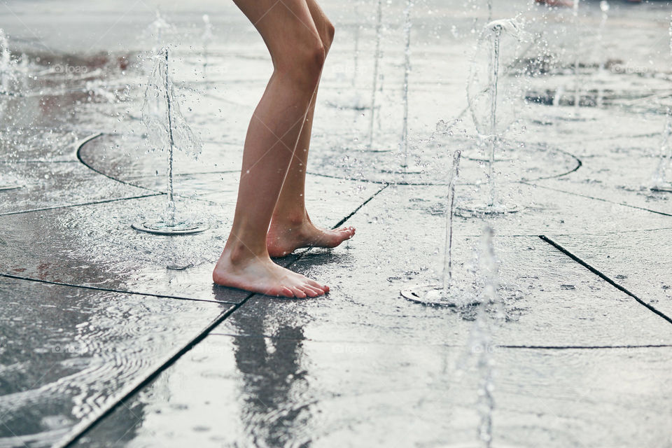 Child standing, jumping, playing in a fountain while hot weather. Closeup of legs, water streams, splashes. Candid people, real moments, authentic situations