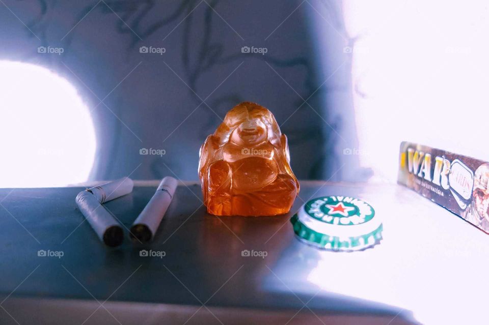 Little orange Buddha statue, with two cigarettes, beer bottle caps, Heineken, and War Game, with distinct lighting, detail shot.