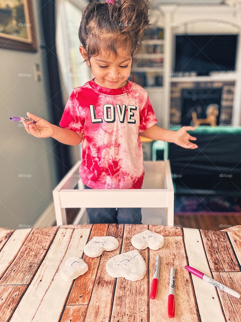 Toddler showing emotion of happiness, child smiling with happiness, toddler is smiling and proud of herself, child making heart Valentines Day gifts, child smiling while showing off work, child making crafts for Valentine’s Day, toddler showing off