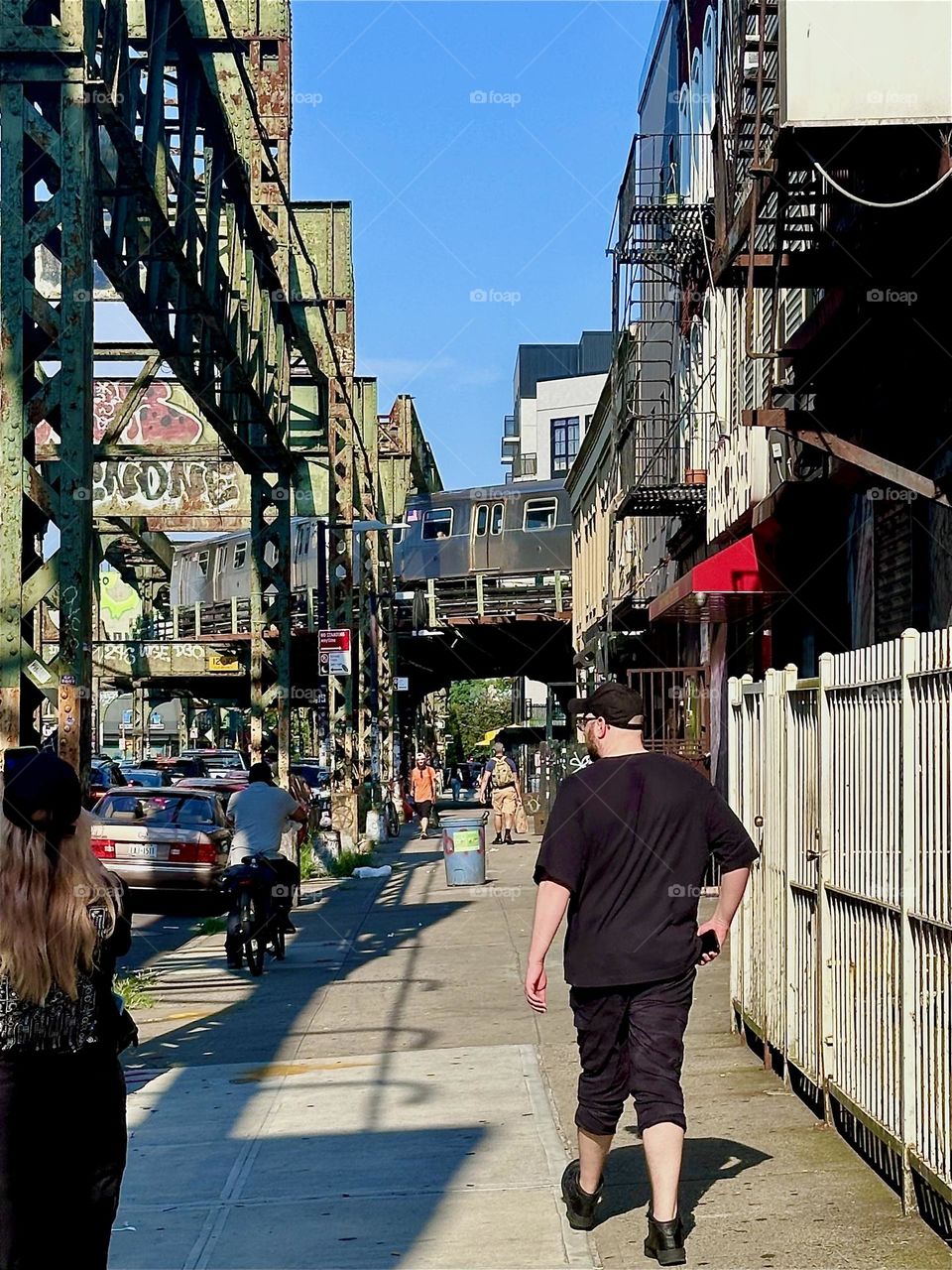 The gentleman is walking down “Myrtle Avenue” in “Bushwick”, Brooklyn along a no longer used early industrial overpass. At the end of the block the “Broadway” overpass turns the corner with the “M” train zooming past. 2024. Hypnotic Productions