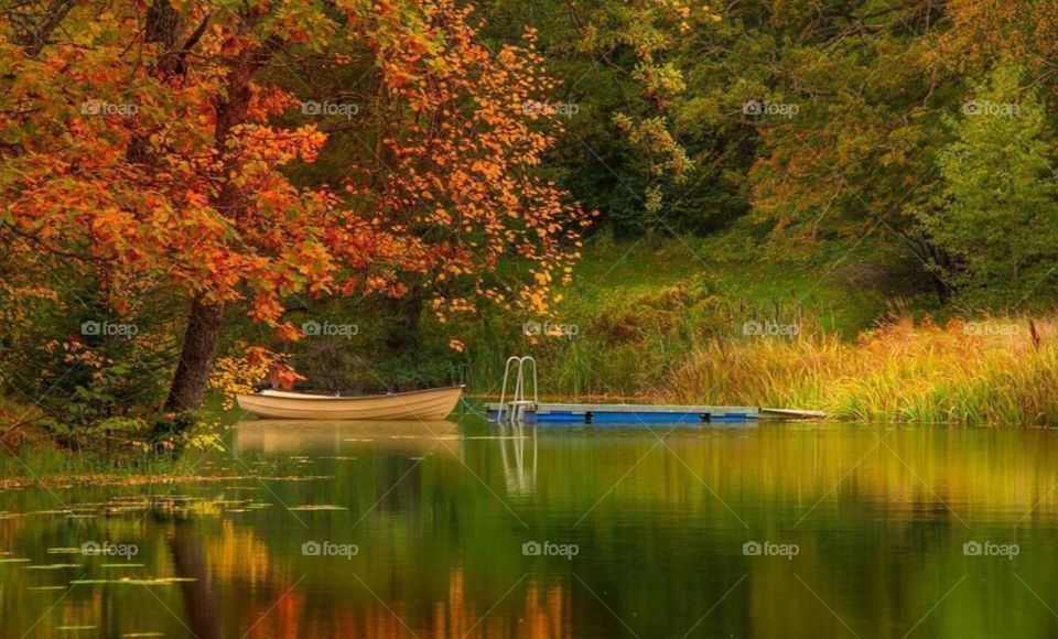 A small wooden boat on a still lake, surrounded by autumn foliage. The trees are reflected in the calm water, their vibrant red, orange, and yellow leaves creating a dazzling display of color.