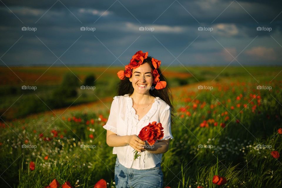 Young millennial woman, being happy and smiling, while being in a poppy field, at sunset on a beautiful summer evening.