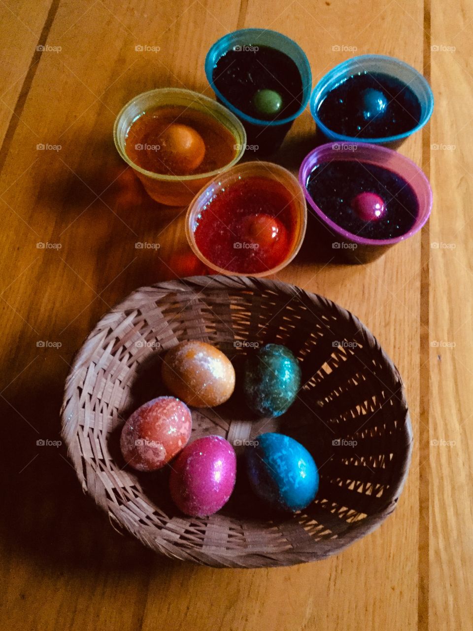 Brightly colored Easter eggs sitting in a basket next to colored eggs sitting cups of dye.