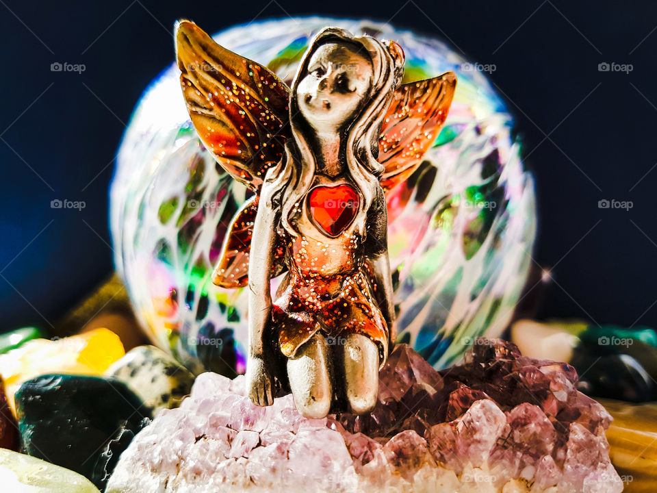 Fairy Sitting on Amethyst surrounded by gem stones with colourful glass ball in background.