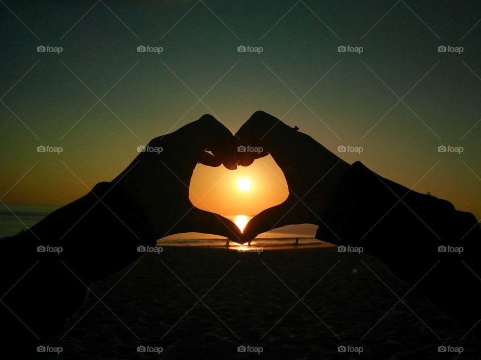 An ocean sunset with a hand of a girl and a hand of a guy making a heartshape around the sun going down in the sea.