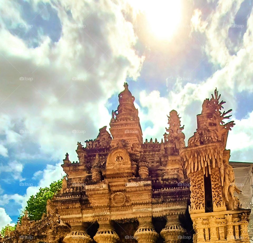 Sculpted Majesty Under the Sun.
Impressive architectural structure with detailed carvings, rising majestically against a bright sky. The sun shines through the clouds, casting a radiant glow over the intricate designs.
