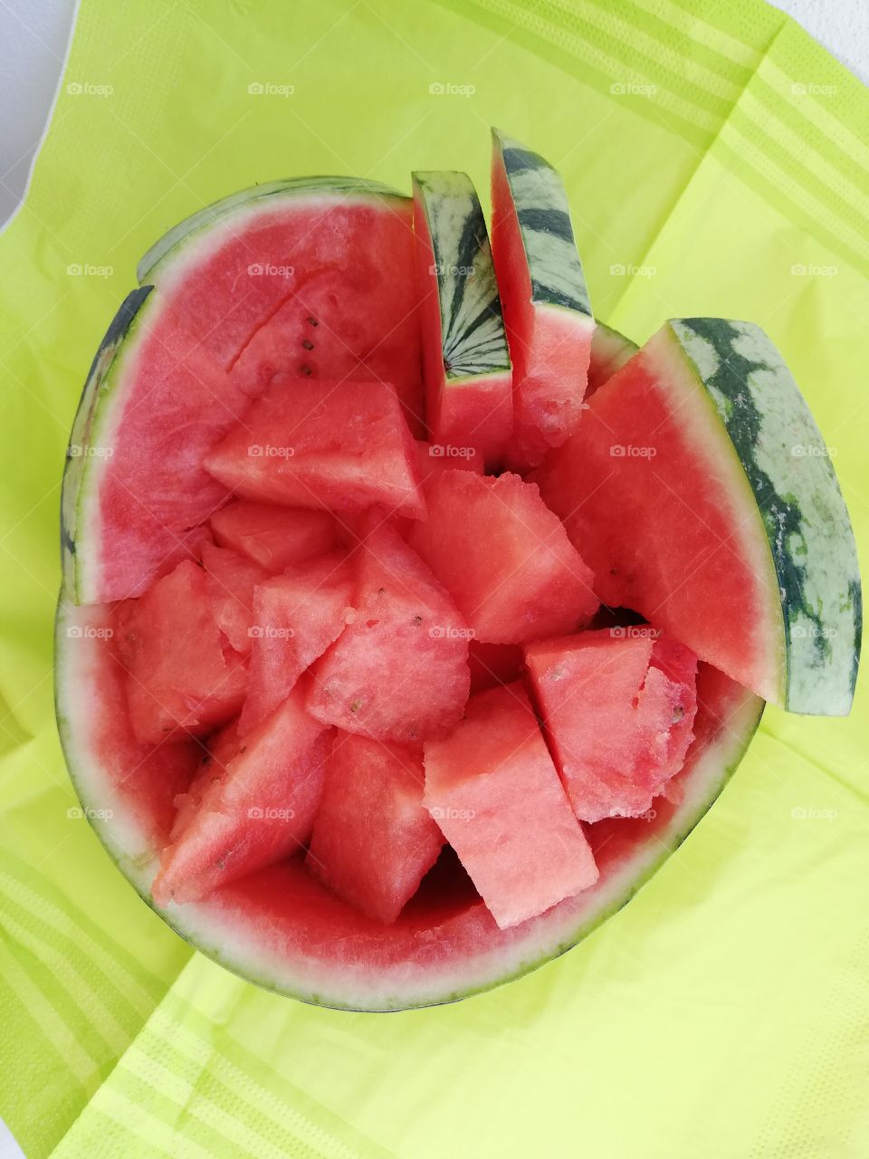 Half of a red juicy watermelon is full of sweet melon cubes and slices seeds inside. It is on the green striped napkin, which is wrinkled.
