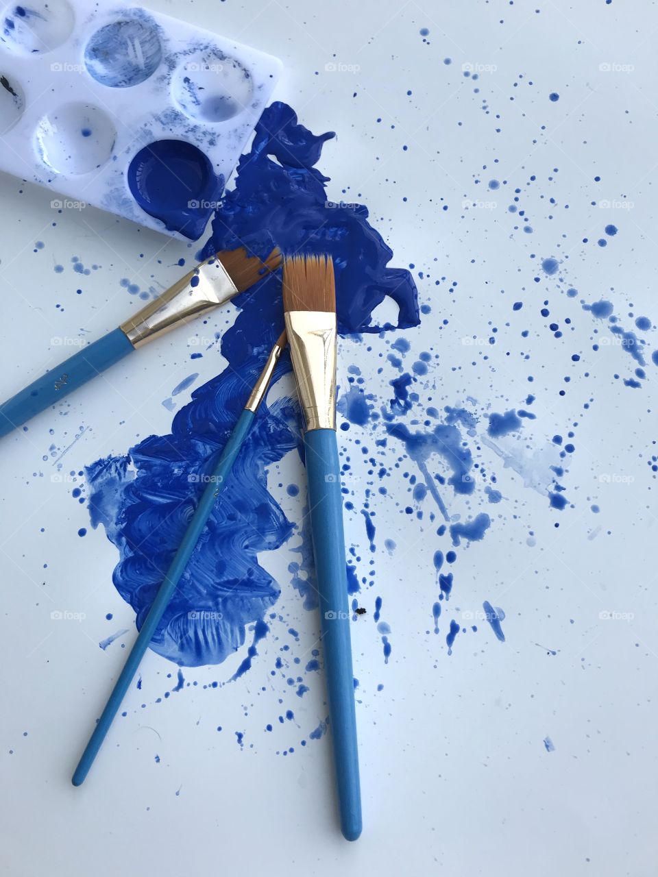 A splattering of blue paint on a piece of white card with three light blue paint brushes!