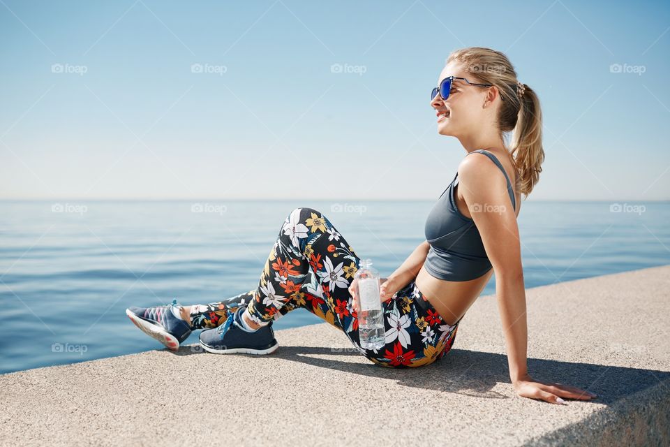 Young fitness blonde woman hold bottle water after fit at beach. Woman sport runner resting taking a break with water bottle drink outside after training.