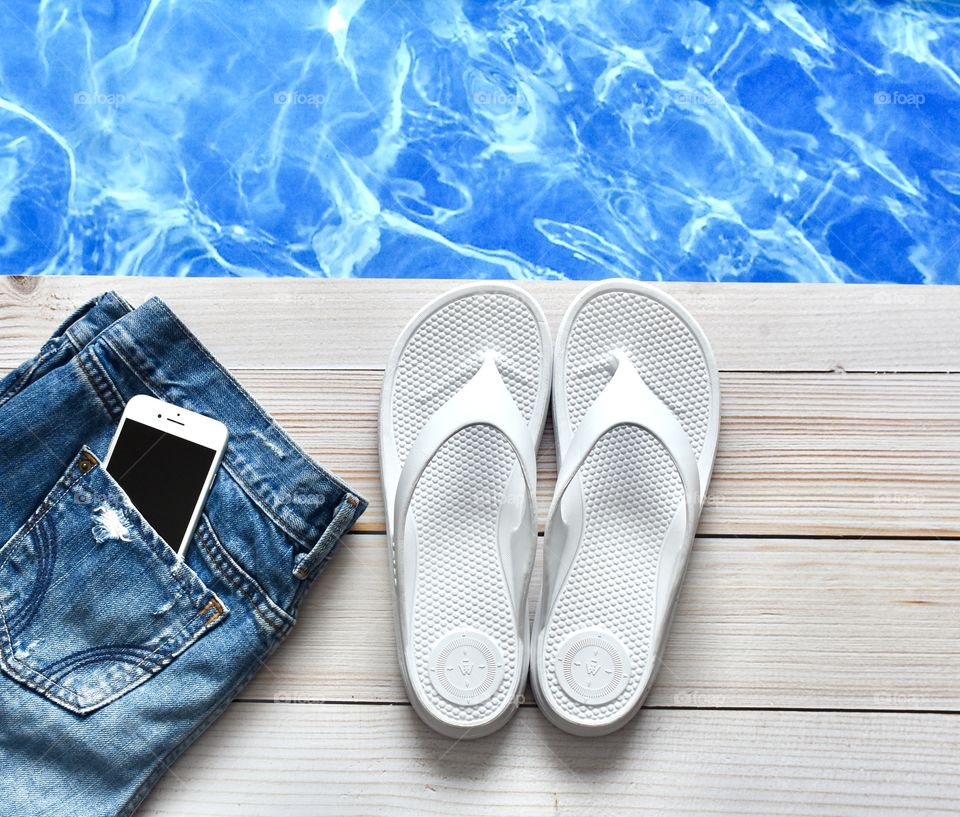 Flat lay of cellphone in Jean shorts pocket with flip flops beside a swimming pool still life 