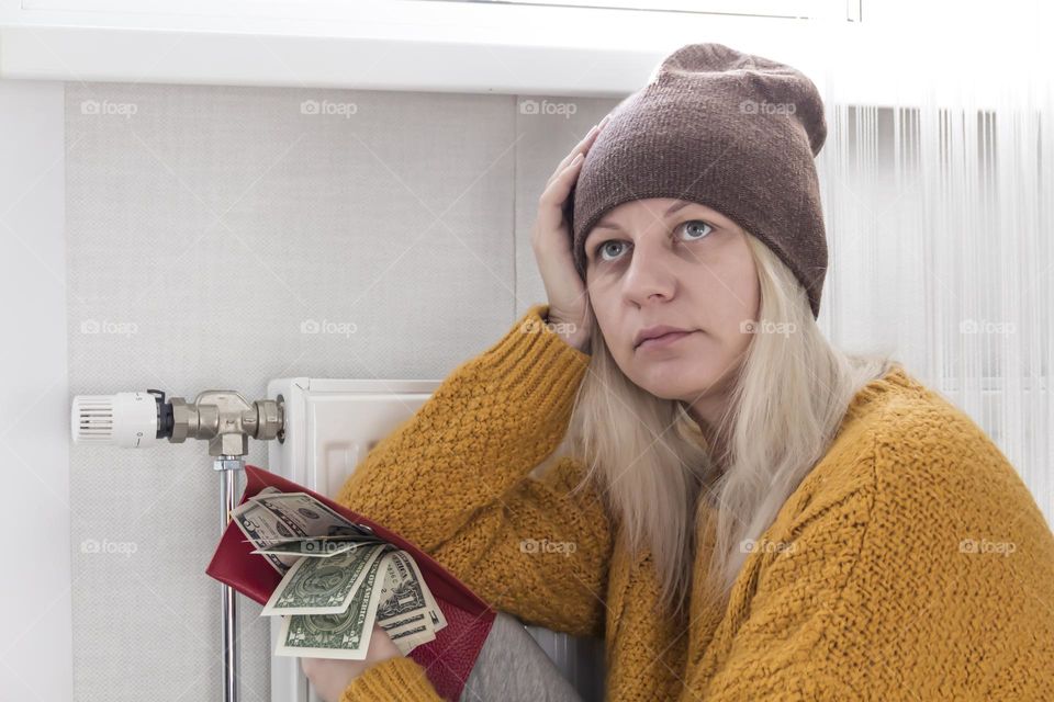 A young girl in a yellow sweater and a brown hat is sitting on the floor, counting money and thinking how to pay bills and taxes near a heater with a thermostat..