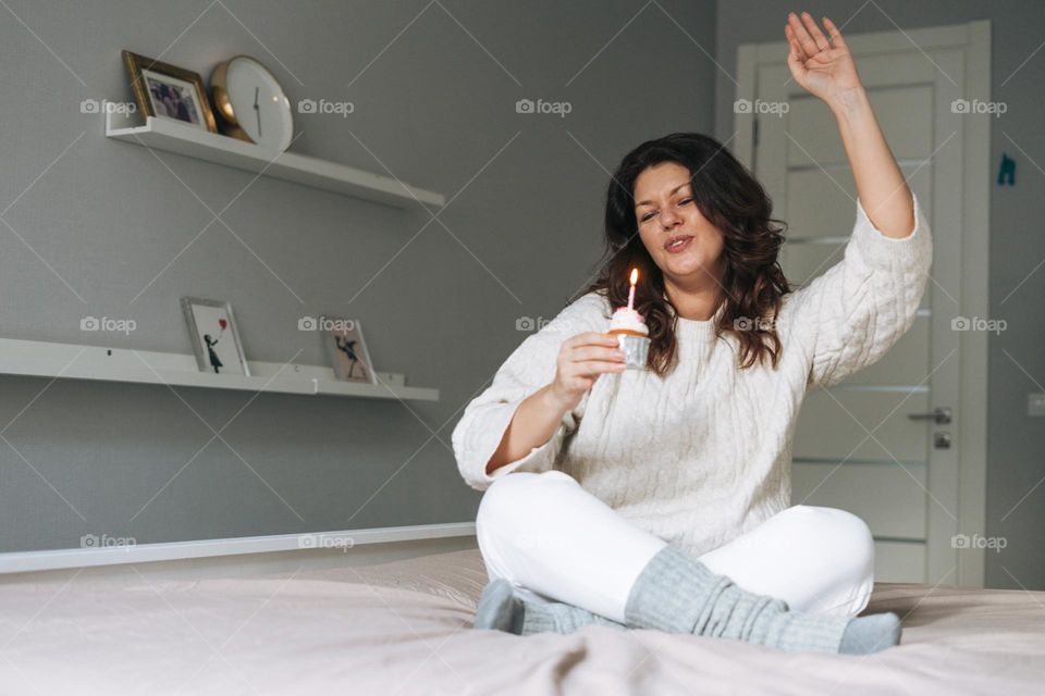 Young adult happy beautiful woman forty years plus size body positive in white cozy knitted sweater with festive cupcake with candle in hands on bed at home party, happy new year, birthday girl
