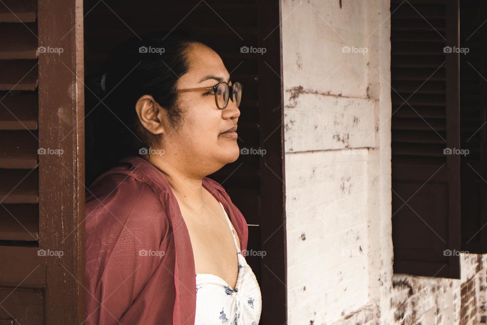 Asian woman with eyeglasses standing near outside the windows of an old asian house facing on the right side.