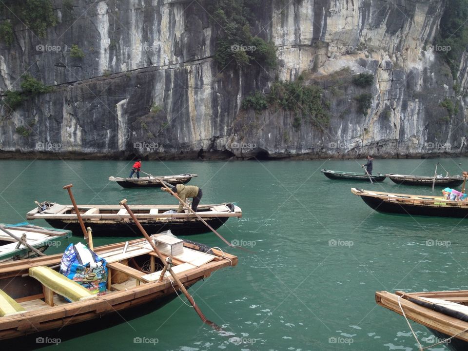 boats and boatman await guests on the blue waters of the bay.The boats are made of wood, with 2 paddles and inside there are wooden bars passing by so that guests can sit on it. in the foreground is very high cliff, gray and grows a few trees