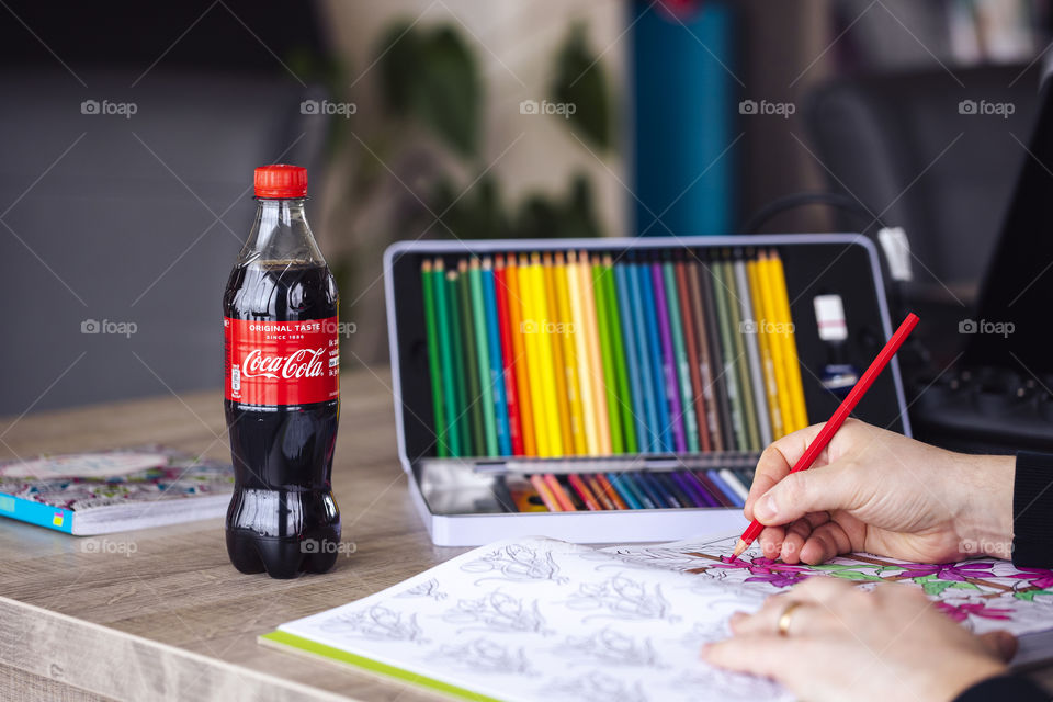 A portrait of someone coloring in a coloring book for adults next to a plastic bottle of coca cola.