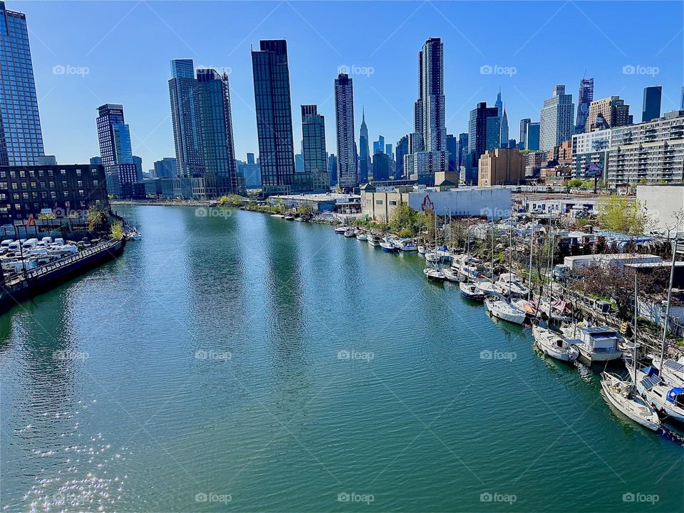 This is “Newtown Creek” with its great variety of boats seen from the “Pulaski Bridge” that connects LIC, Queens to “Greenpoint”, Bklyn. Across the “E River” in the distance we see “Manhattan” incl. the “Empire State Bldg”. 2024. Hypnotic Productions