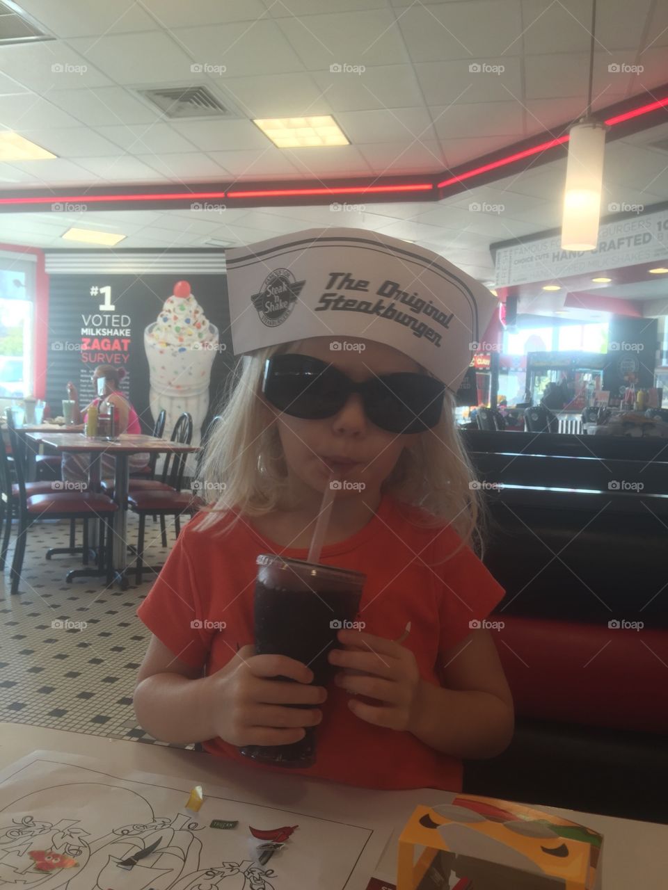 Cool girl drinking a soda pop. Toddler at diner wearing a funny hat and sunglasses
