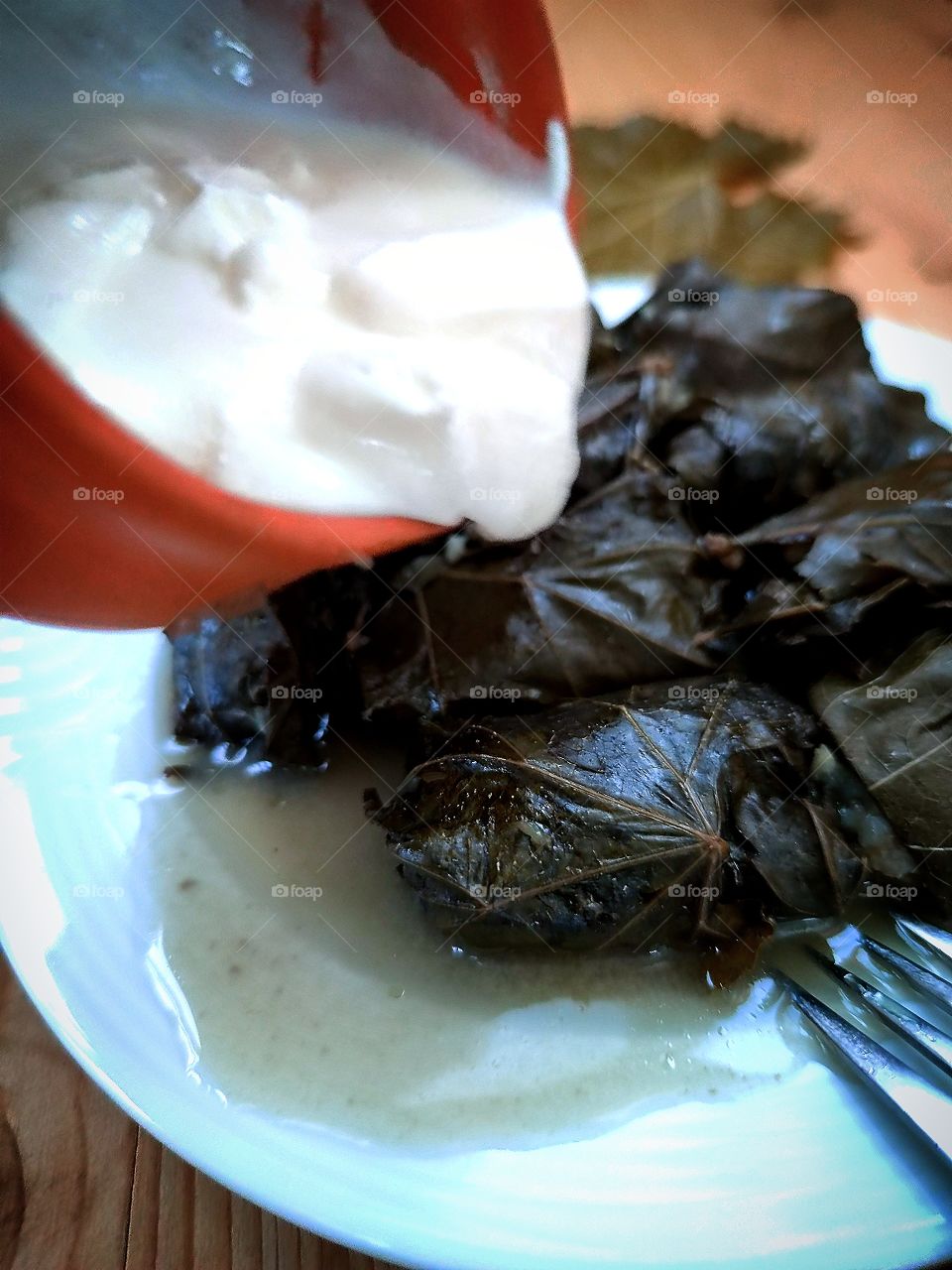 Food.  Dolma on a plate.  Fork tines.  Clay gravy boat with sour cream over dolma.  Dolma is minced meat wrapped in grape leaves