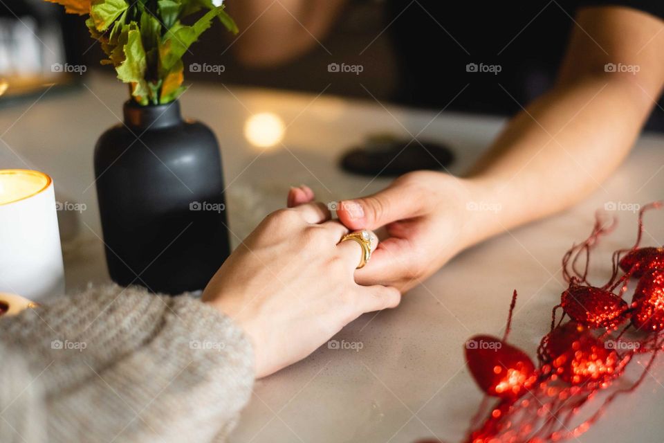 They reach across the table and hug each other.
On the table are red hearts, a black vase with green stems, and a light candle.