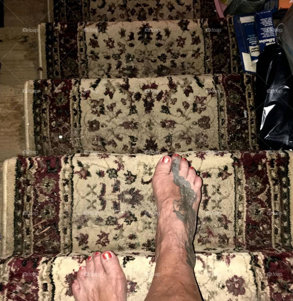 Bare feet walking down stairs. These are womens feet with painted toenails stepping carefully. Each foot has a tattoo artistically done.  Stairs are covered with an oriental runner.