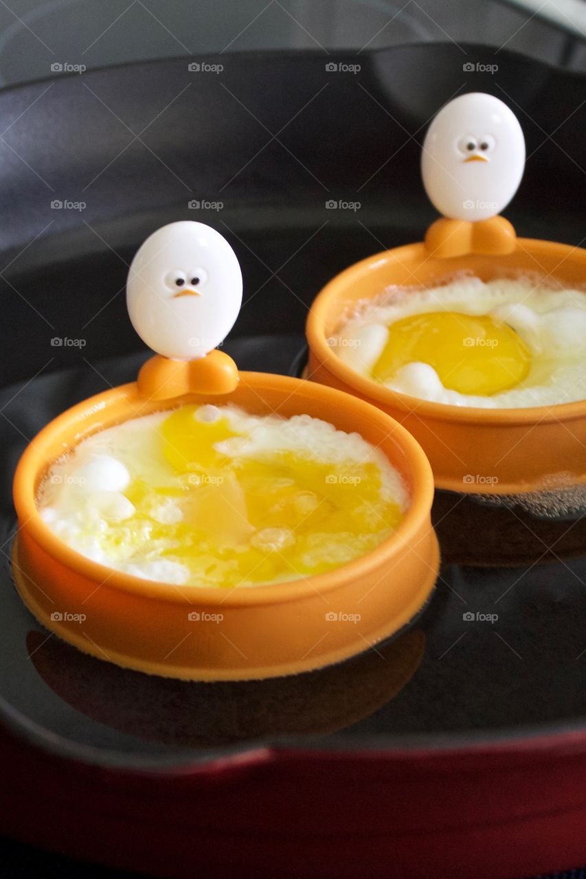 Closeup of a cast iron pan containing eggs being fried in silicone egg rings with egg-shaped handles depicting baby chick faces and feet