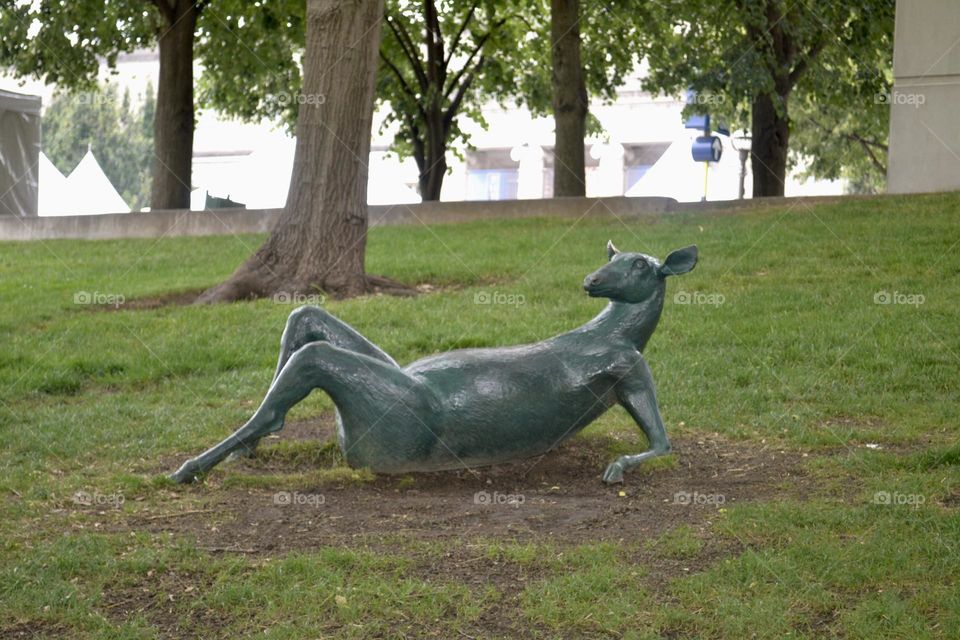 A green sculpture of a deer lying on its back propped up on its elbows and bent knees on brown dirt and green grass with trees nearby
