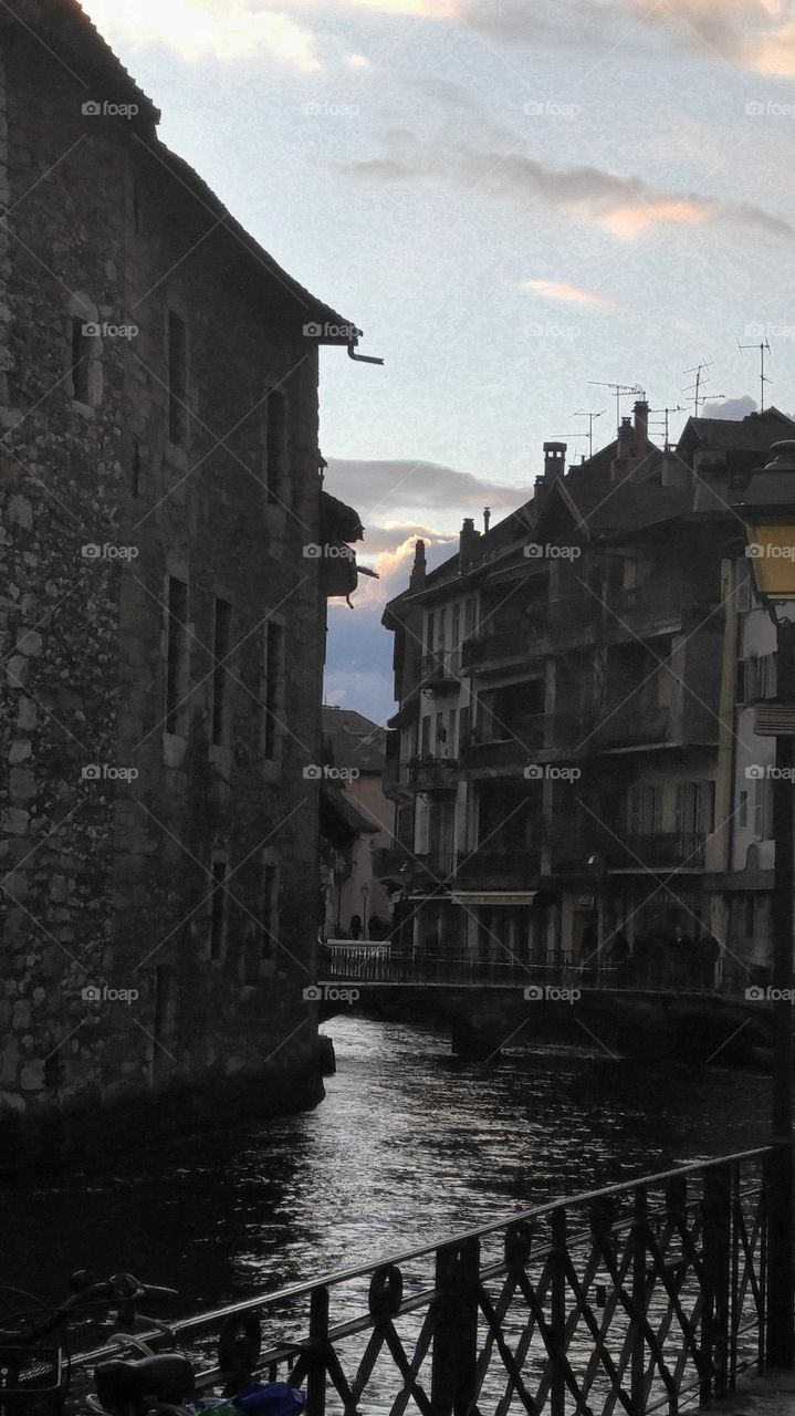 Sunset through shadowy, old buildings with a bridge over water.
