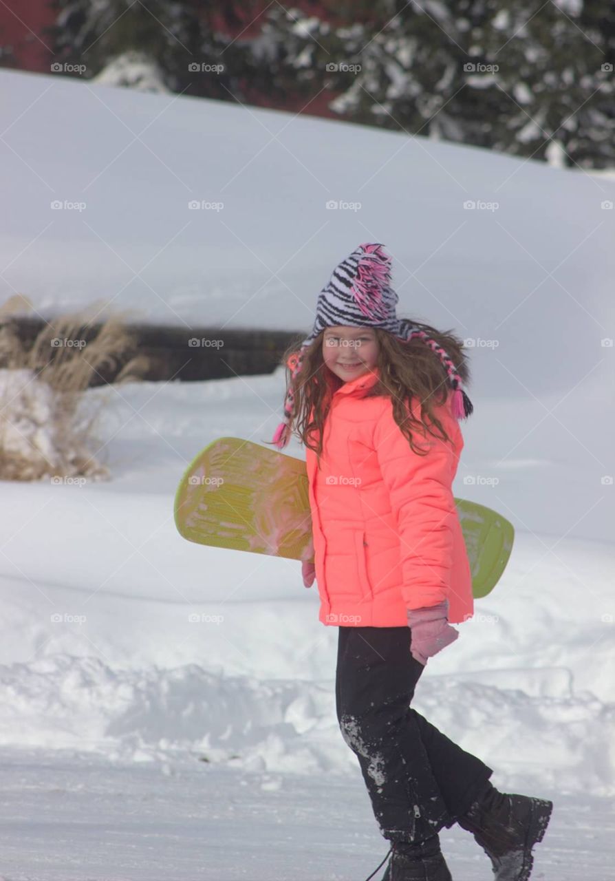 A nine-year-old little girl, all bundled up in her winter gear, carrying her snowboard up the hill.
