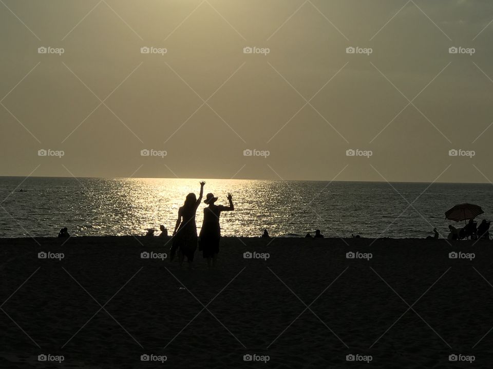 Two women in silhouette waving at camera during dusk with a calm sea behind them 