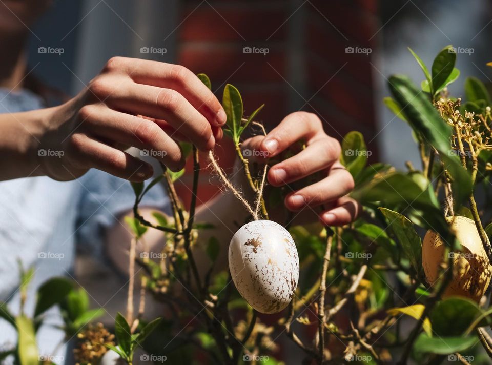 The hands of a caucasian teenager girl hang a white marble decorative easter egg with a jute thread on a green shrub growing in the backyard of the house on a clear sunny day, close-up side view. Easter tradition concept.