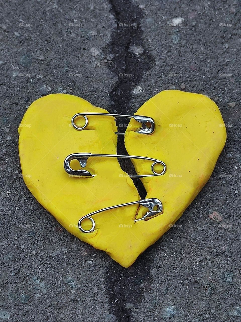 Yellow. A yellow heart with a crack lies on a crack on the asphalt. Cracked heart chipped with metal pins