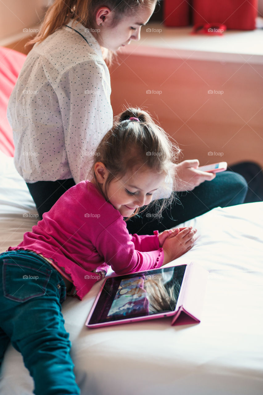 Teenage girl using mobile phone together with her little sister watching animated movie on tablet, both girls sitting in bed in bedroom