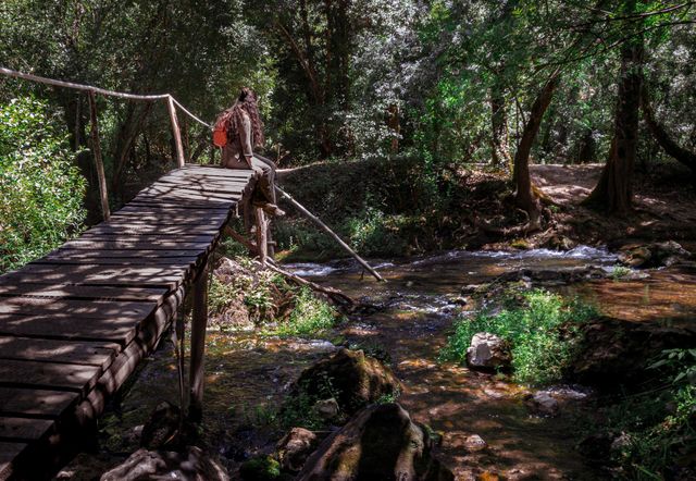 A hiker takes a break sitting on a bridge over a small stream in woodland