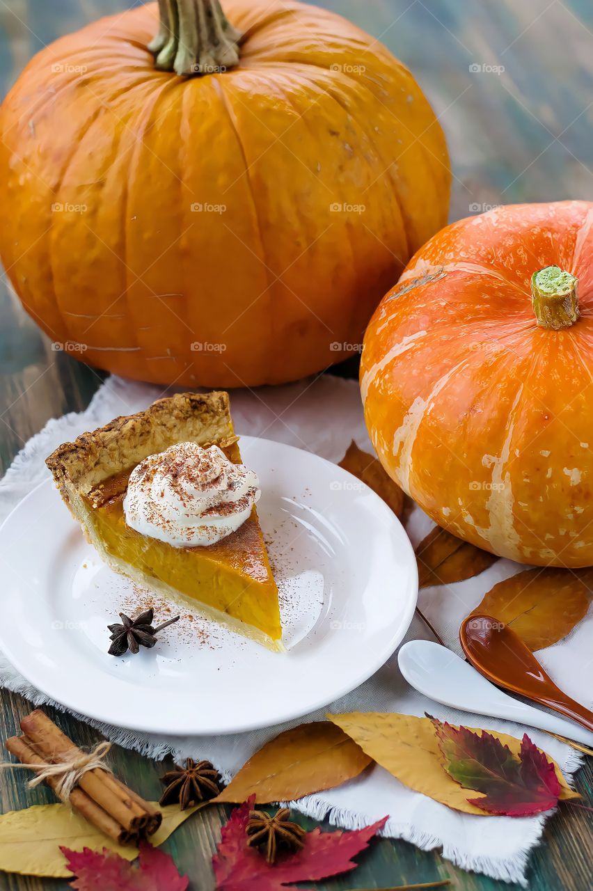 Close up of a slice of pumpkin pie on white plate alongside two bright orange pumpkins.