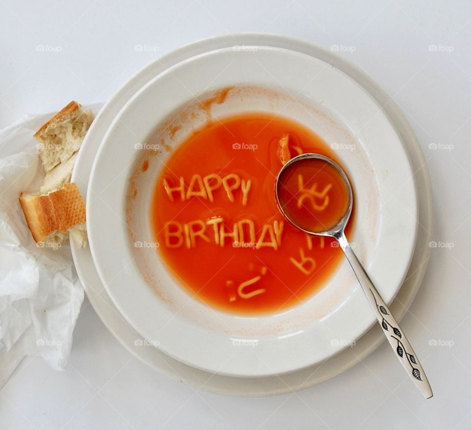 A bowl of soup with pasta letters spelling out the message happy birthday, with white bowl, silver spoon and bread roll. 