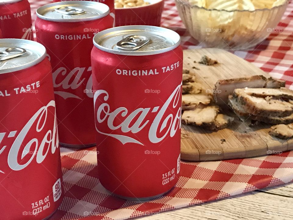 Cans of Coca-Cola on a red and white checkered background with barbecue food and potato chips 