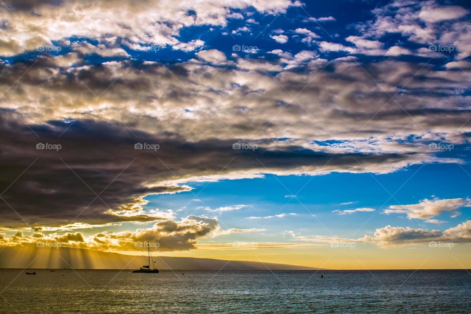 Lone sailboat sailing off into the sunset on the beautiful calm waters of the Hawaiian Islands with stunning clouds and rays of sunshine peering through 