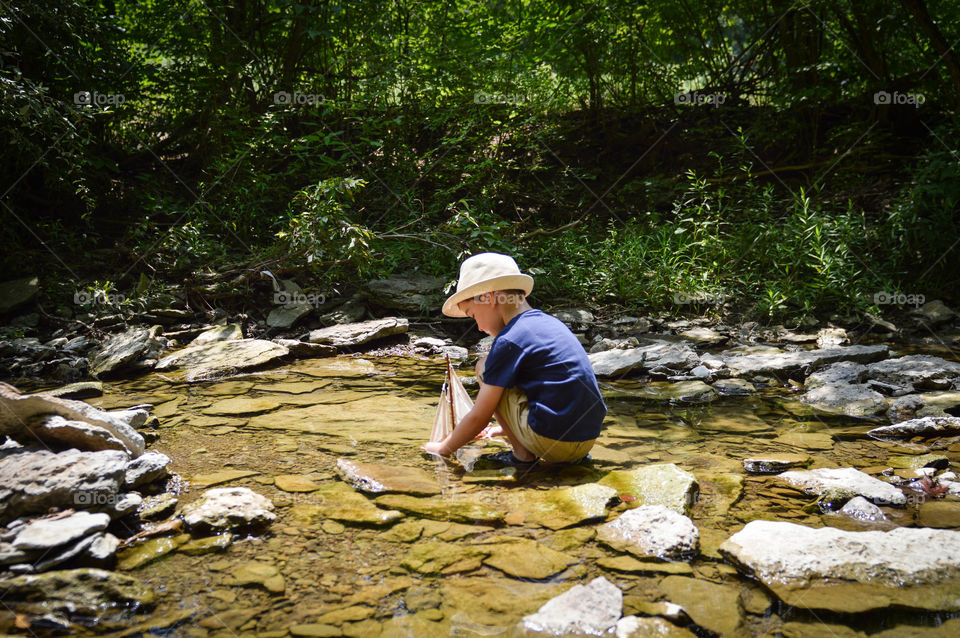 Boy playing with a toy sailboat in a creek during the summer