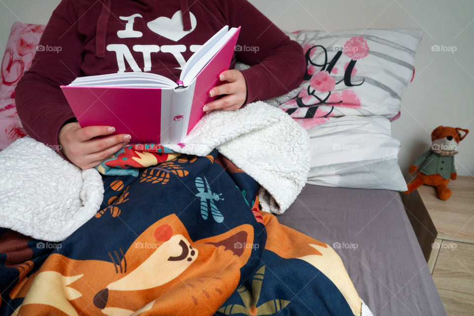 A young girl is reading a book in bed wrapped in a comfortable warm blanket.