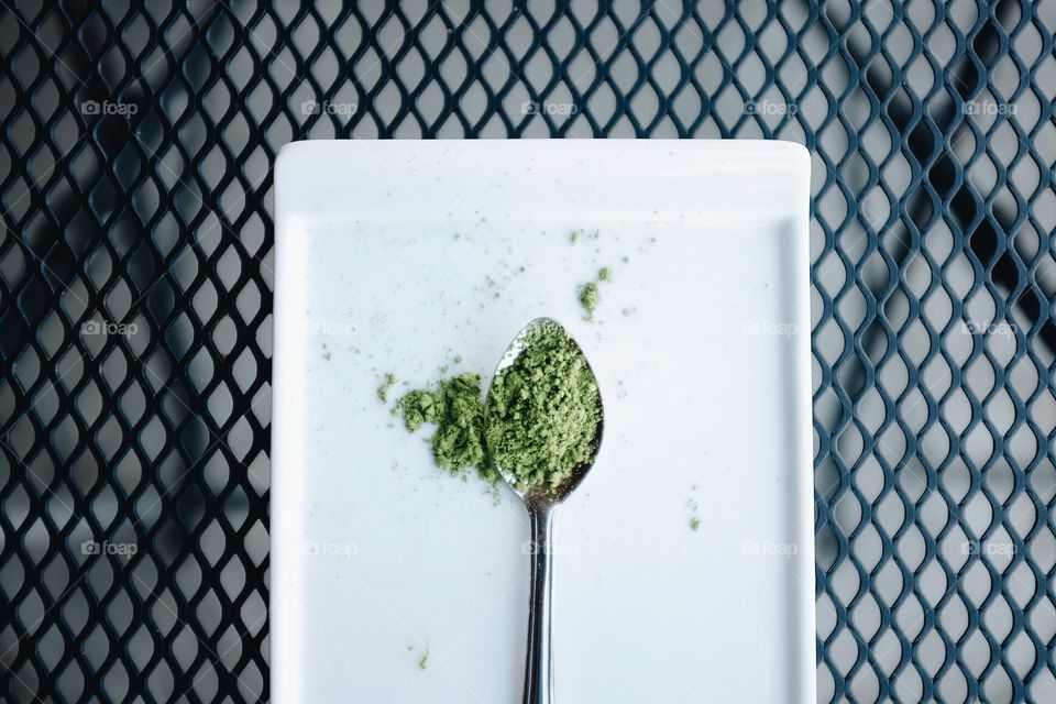 Green matcha tea powder on a small spoon sitting on a white plate that is sitting on a wire mesh table. 