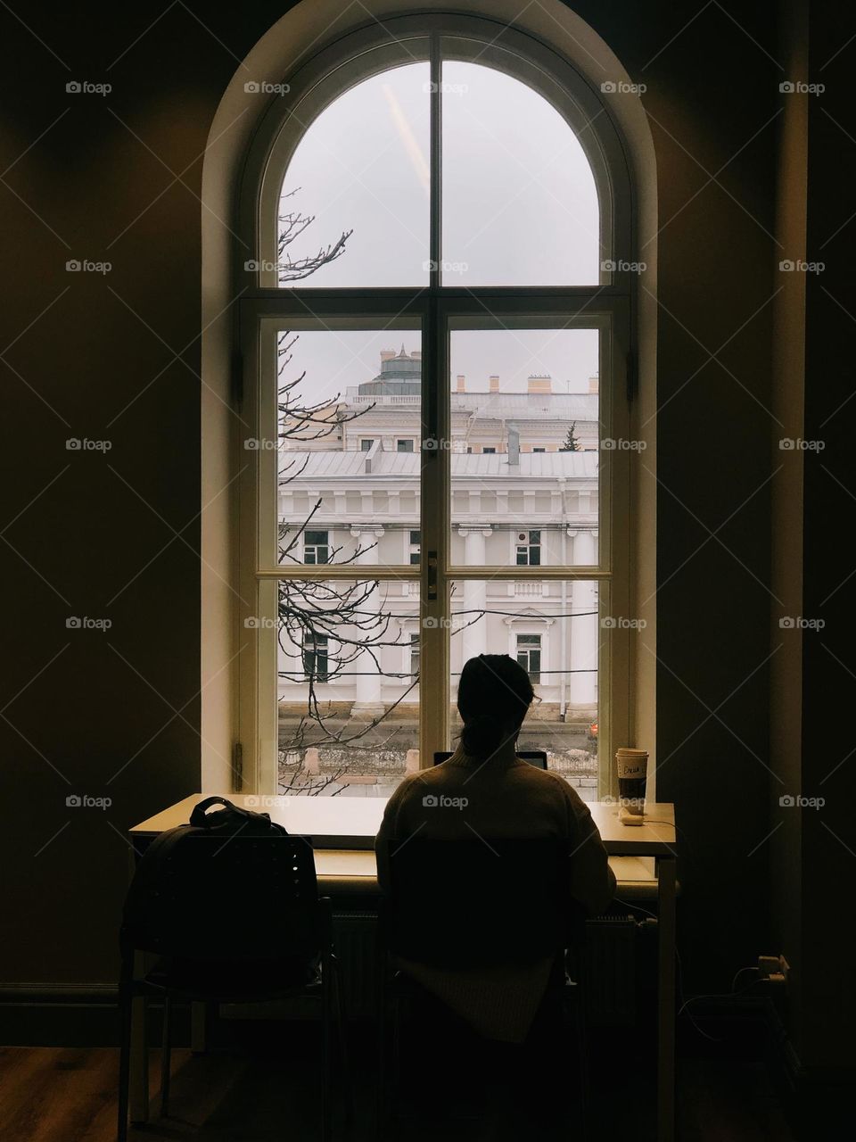 A young girl sits at a table in the library by the window, a silhouette of a girl, back view.