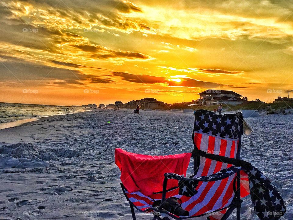 A lonely red, white, and blue chair abandoned at the beach on a stunning sunset