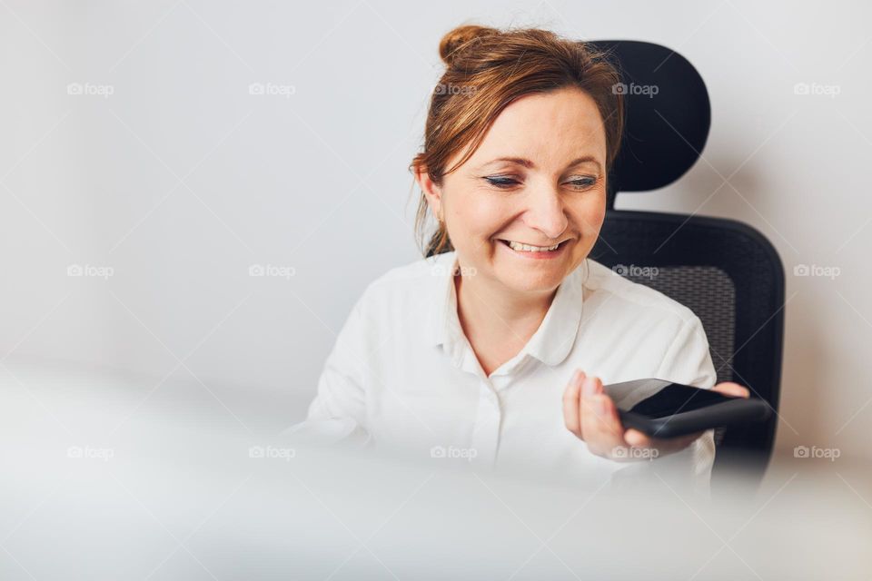 Businesswoman talking on mobile phone using speaker mode. Woman recording audio message using voice assistance and recognition function on smartphone. Female holding smartphone sitting in chair in office