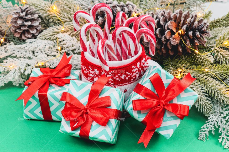 Three Gift Boxes with a red ribbon, lollipops in a mug with a Christmas sweater, a fir branch and a burning garland on a green background, close-up side view. Holiday creative gifts concept.